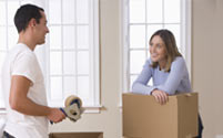 A man holding a packing tape dispenser looks at a woman leaning on a box.